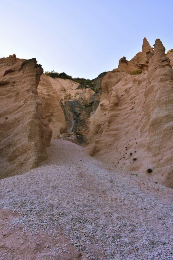 Lame Rosse hike. A view from inside a narrow canyon at Lame Rosse in the Marche region, featuring towering, sharp-peaked rock formations and eroded earth pinnacles on either side of a steep, grey gravel path leading upward under a pale blue sky.