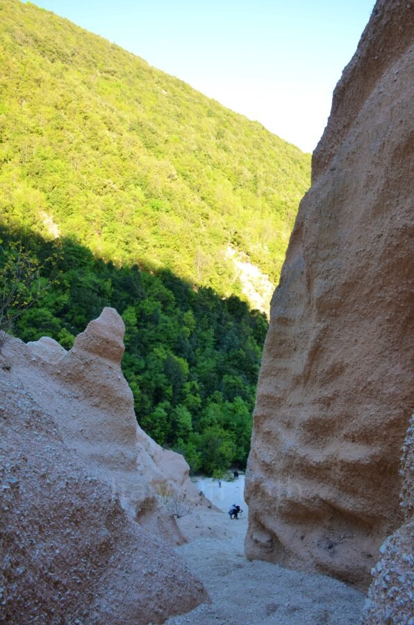 A steep downward view from the top of the Lame Rosse canyon, showing the narrow gravel descent between tall, textured red rock pinnacles, with tiny figures of people visible at the bottom and a densely forested green hill rising in the background.