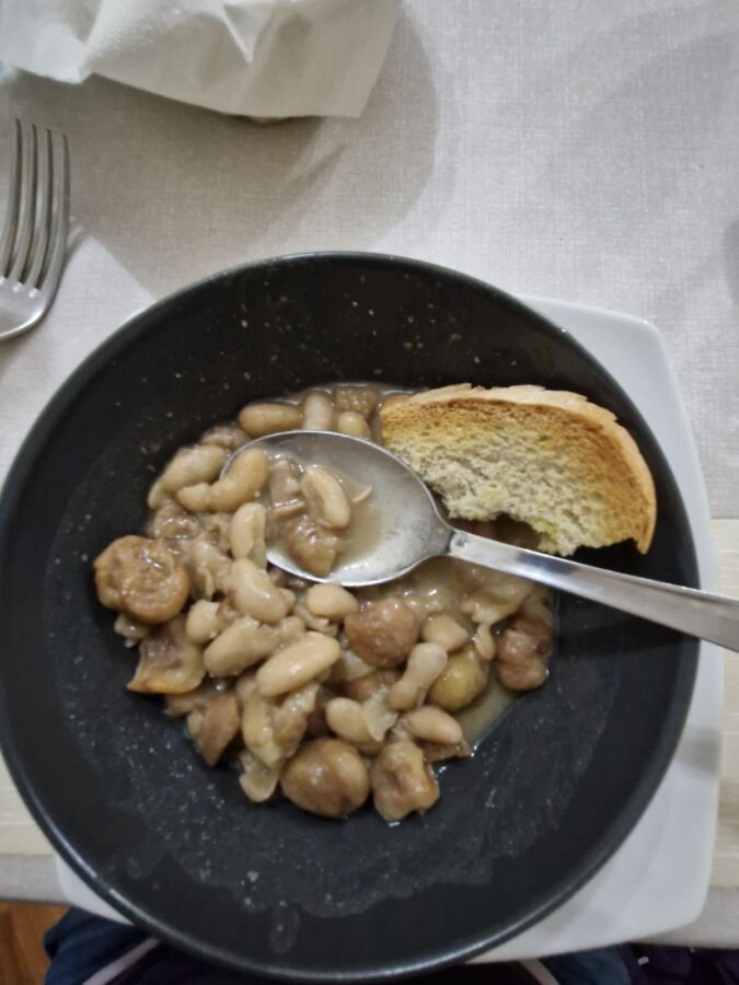 A bowl of white bean and chestnut soup in a dark bowl, served with a slice of toasted bread and a metal spoon. The dish is photographed from above on a light-colored tablecloth.