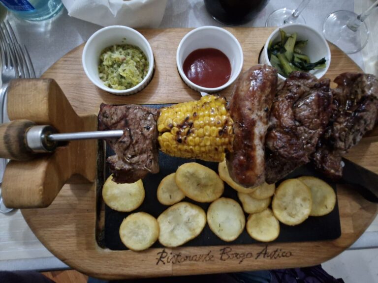 Monti Azzurri Montemonaco. A rustic meat platter served on a wooden board at Ristorante Borgo Antico, featuring a metal skewer with grilled beef, sausage, and corn on the cob over a bed of roasted potato slices, accompanied by three small white bowls containing sauces and cooked chicory