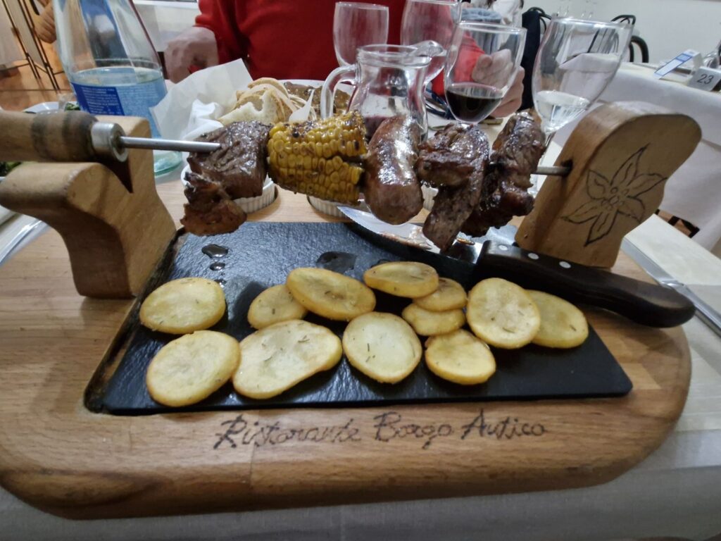 A metal skewer with grilled meat, a corn cob, and sausage suspended over a black slate board with roasted potato slices. The serving set is mounted on a wooden base engraved with "Ristorante Borgo Antico," placed on a restaurant table with wine glasses and bread in the background.