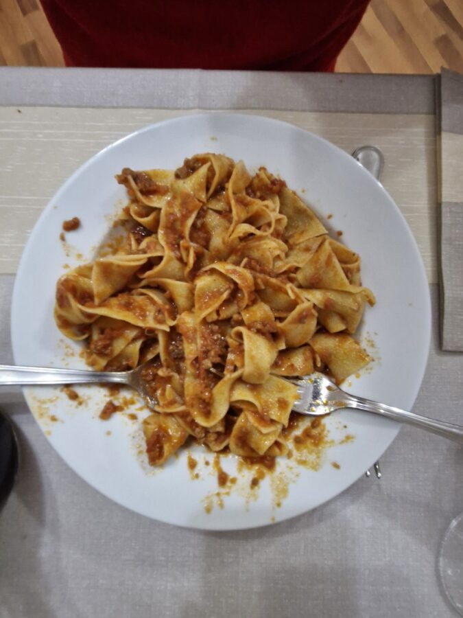 A top-down view of a plate of wide ribbon pasta coated in a thick, rustic wild boar ragu, served at Ristorante Borgo Antico with two forks resting on the sides of the white plate.