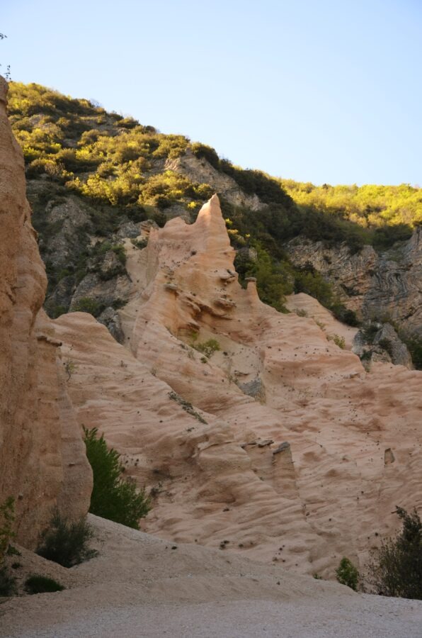 A vertical low-angle shot of the towering red-orange rock pinnacles and layered cliffs of Lame Rosse against a clear blue sky, with a gravel slope in the foreground and green trees on the right.