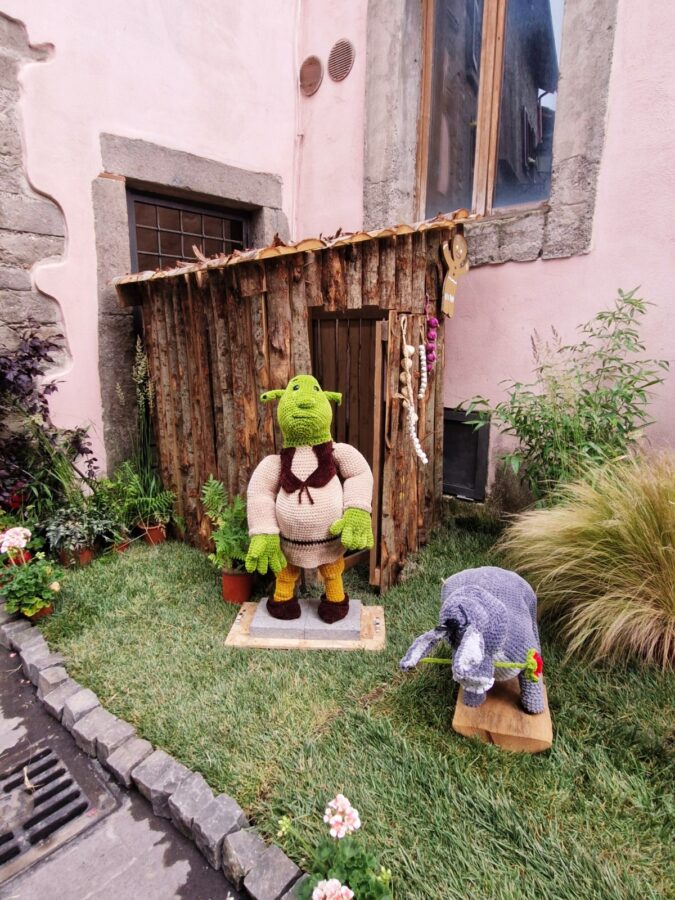A life-sized, crocheted Shrek figure stands on a small patch of grass in front of a rustic wooden shack made of logs. Next to him, a smaller crocheted Donkey figure holds a red rose in its mouth. The display is set up against a pink building wall with surrounding potted plants during the Peperino in Fiore festival.