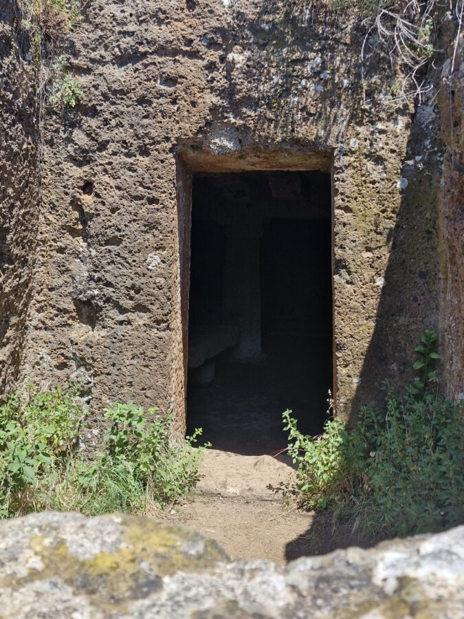 The rectangular stone entrance of Tomba Margaretha cut into a volcanic tufa rock wall with small green plants growing at the base in Valle Capellana.