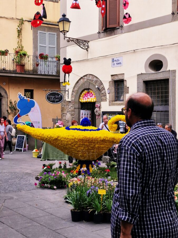A large, yellow and blue floral display shaped like Aladdin's magic lamp stands in a small stone square called Piazza S.S. Trinità during the Peperino in Fiore festival. A blue cardboard cutout of the Genie rises from the spout of the lamp. A man in a blue plaid shirt stands in the foreground with his back to the camera, looking at the display, while red and black Mickey Mouse decorations hang overhead.