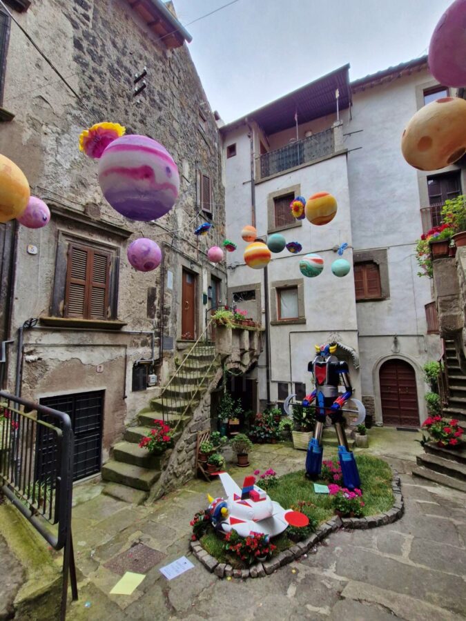 Multiple colorful, painted planet models hang suspended in the air between old stone residential buildings in a narrow courtyard. On the ground below, a large toy robot figure stands on a small patch of grass next to a spaceship model surrounded by red flowers during the Peperino in Fiore festival.
