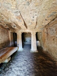 Hikes near Rome. The dark interior of the ancient Etruscan Tomba Margaretha, showing two hand-carved stone columns supporting a rock ceiling with carved beams and funeral stone platforms.