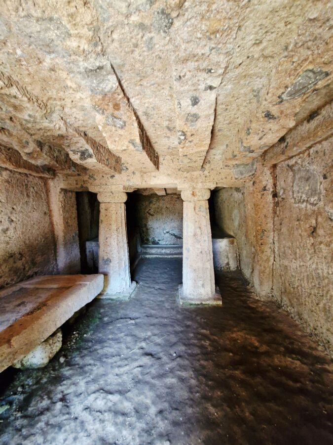 Hikes near Rome. The dark interior of the ancient Etruscan Tomba Margaretha, showing two hand-carved stone columns supporting a rock ceiling with carved beams and funeral stone platforms.
