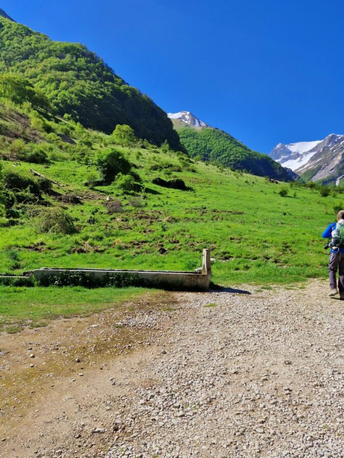 A stone water fountain and a long concrete trough sit next to a gravel path in a wide valley. Bright green, grassy hills rise up toward distant mountain peaks covered in patches of snow under a clear blue sky. A hiker with a backpack walks away along the trail on the rig