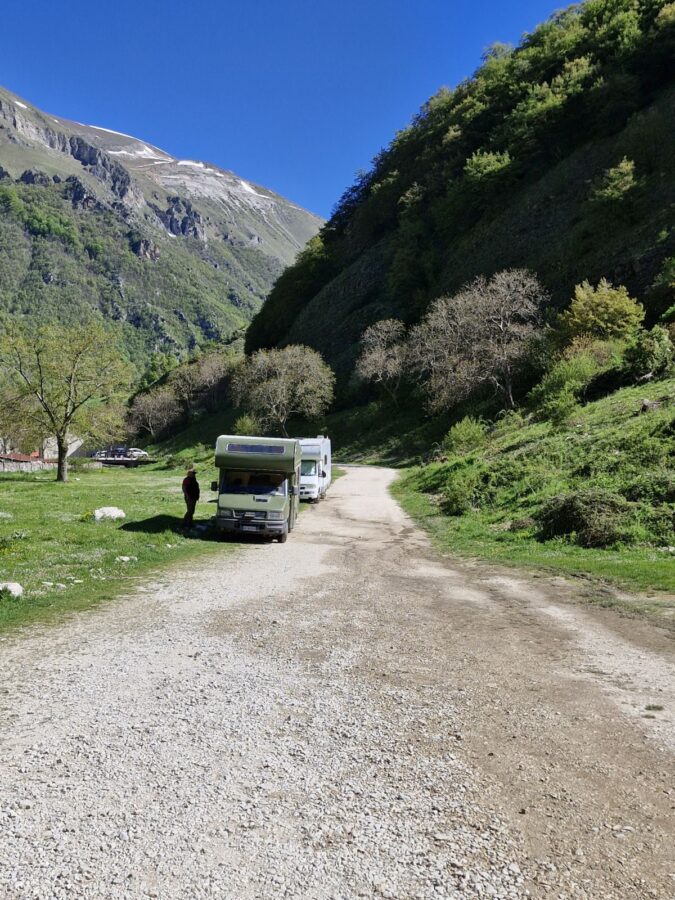 A wide gravel road leading into a mountain valley with steep green hills and a clear blue sky. Two camper vans are parked on the side of the track next to a grassy area, with a person standing nearby.