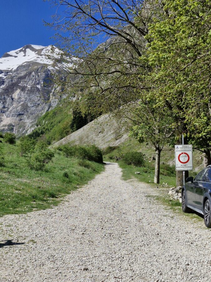 A wide gravel hiking trail leading into a mountain valley under a deep blue sky. Green grass and leafy trees line the path, with a partially snow-covered peak rising sharply in the background. A white road sign with a red circle and part of a dark grey car are visible on the right.