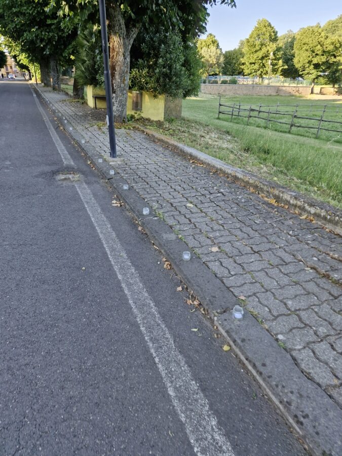 A close-up view of numerous small glass jars containing tealight candles arranged in rows along an asphalt street curb in Villa San Giovanni in Tuscia.