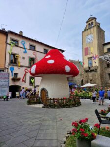 Local festivals in Italy. A large, mushroom-shaped house decoration resembling a red and white fly agaric sits in the center of a stone-paved main square during the Peperino in Fiore festival. The surrounding historic stone buildings are decorated with colorful oversized keys on the walls, and a tall stone clock tower stands in the background under a cloudy sky.