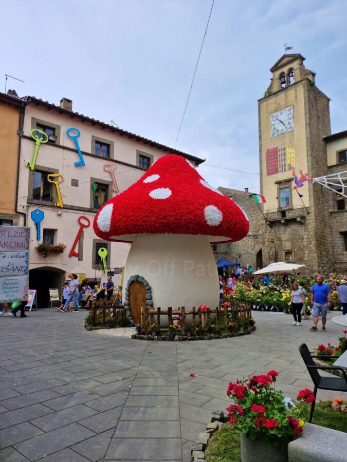 Local festivals in Italy. A large, mushroom-shaped house decoration resembling a red and white fly agaric sits in the center of a stone-paved main square during the Peperino in Fiore festival. The surrounding historic stone buildings are decorated with colorful oversized keys on the walls, and a tall stone clock tower stands in the background under a cloudy sky.