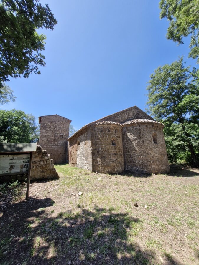 The stone facade of an ancient medieval church standing alongside a dirt path on the Cammino dei Tre Villaggi trail, surrounded by green forest trees and overgrown grass under a blue sky.