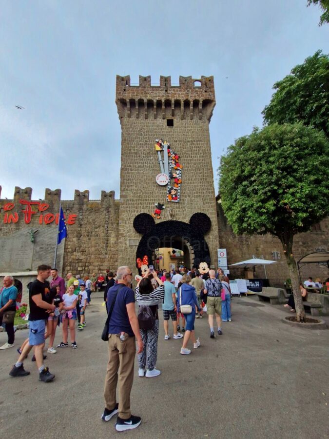 A crowd of people gathers in front of an ancient stone fortress wall and tower gateway decorated for the Peperino in Fiore festival. The archway of the stone tower is framed by a massive black floral display shaped like Mickey Mouse ears, flanked by Minnie and Mickey cartoon cutouts. A large film-strip decoration with cartoon characters climbs up the tower wall under a cloudy sky.