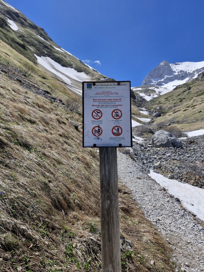 A wooden signpost stands next to a rocky trail in a high mountain valley with melting snow patches under a clear blue sky. The white informational sign lists regulations for a protected area in the Monti Sibillini National Park, showing various red prohibition icons