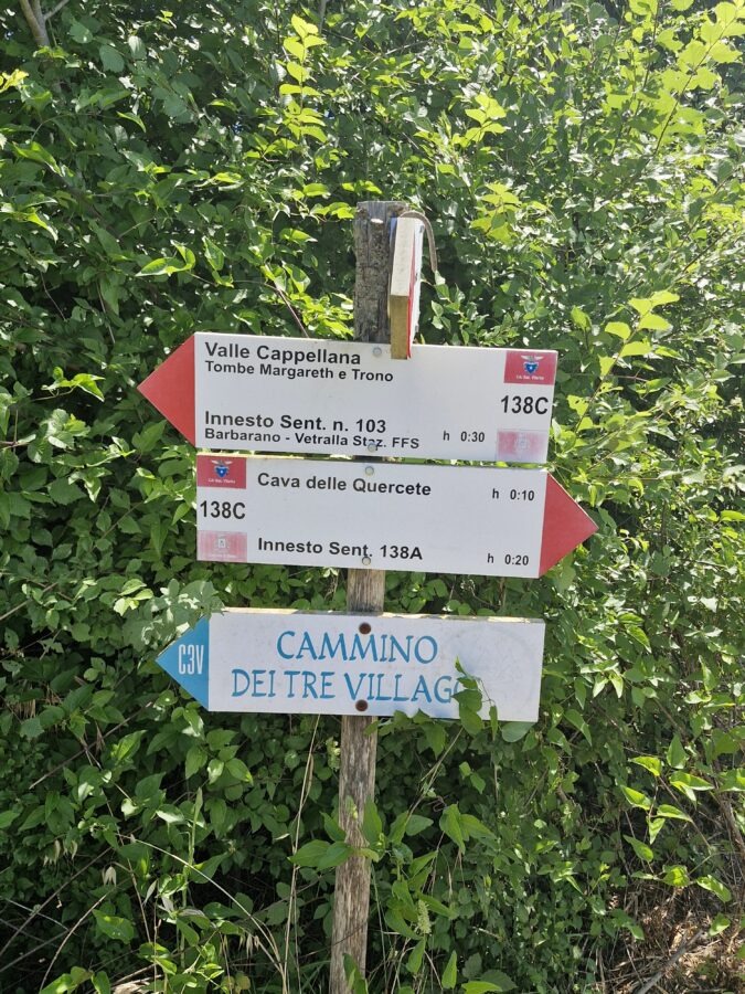 A wooden post with multiple hiking signs, including red-and-white indicators for Valle Cappellana and Cava delle Quercete, above a blue-and-white arrow for the Cammino dei Tre Villaggi trail against green foliage.