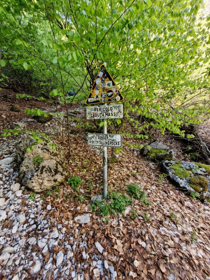 A metal signpost stands on a steep, rocky mountain path covered in dry leaves and surrounded by green trees. The post holds a yellow triangular danger sign covered in stickers, an Italian sign reading Pericolo Caduta Massi, and an English sign reading Danger Risk of Falling Rock