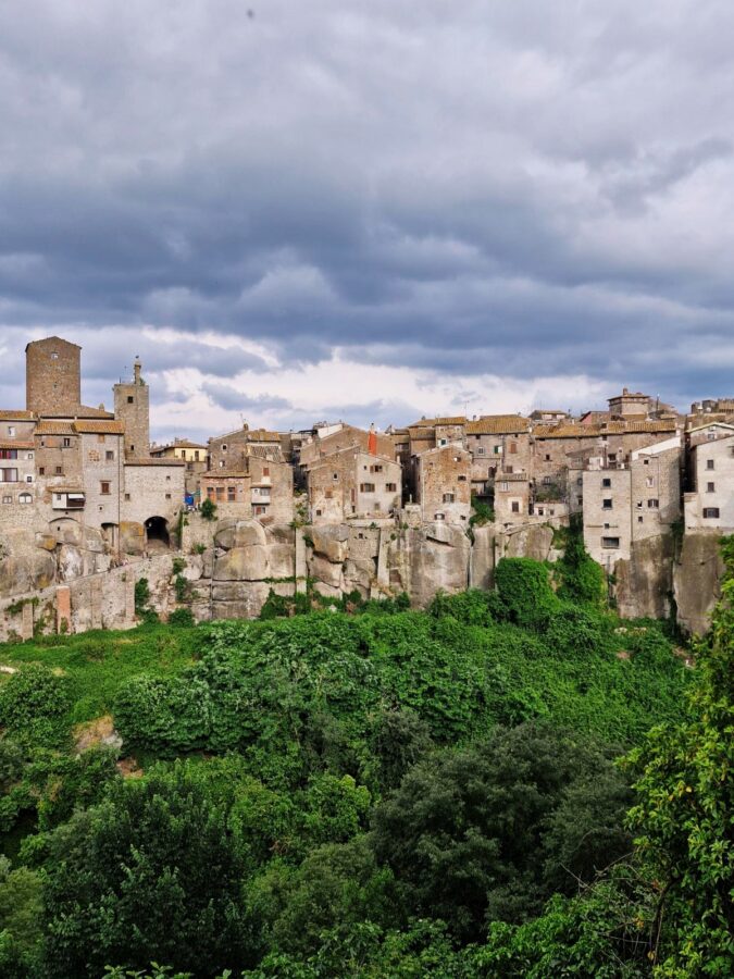 A wide shot of an ancient stone borgo built directly on top of a steep, rocky cliffside. Lush green trees and vegetation cover the valley below the stone houses. A prominent medieval tower and an arched gateway stand on the left side of the village under a heavy, overcast sky filled with dark clouds.