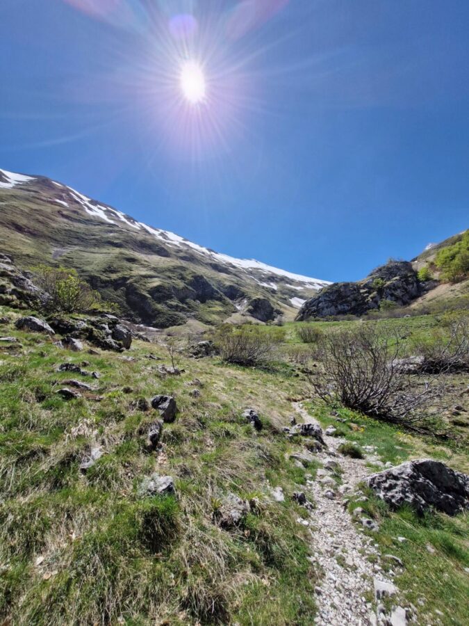 hike to Lago di Pilato from Foce. Narrow, rocky hiking trail cutting through a green mountain valley under a bright sun with prominent lens flare, leading toward snow-patched peaks in the