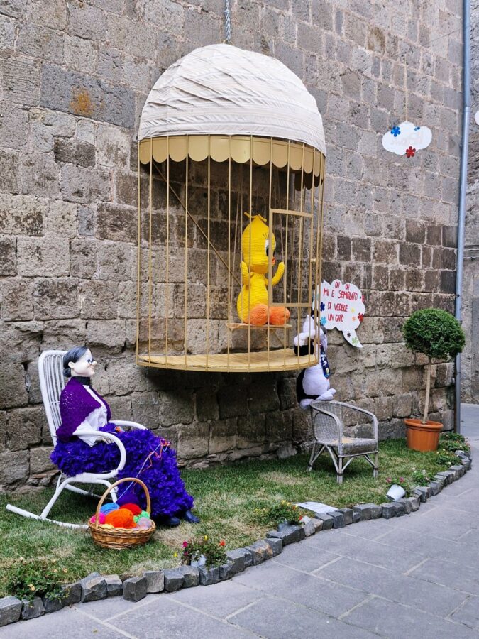 A large, knitted yellow Tweety Bird character sits inside an oversized birdcage hanging against an old stone wall. On a small patch of grass below, a life-sized mannequin of Granny sits in a white rocking chair next to a basket of yarn, while a Sylvester the Cat figure climbs near a small empty chair during the Peperino in Fiore festival.