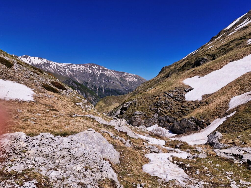 "A rugged high-altitude mountain valley under a clear blue sky. Rocky, grass-patched slopes and fields of melting snow frame a deep gorge, with larger snow-covered peaks visible in the distance