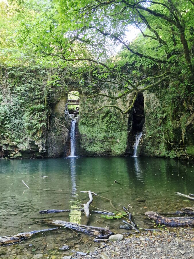A small waterfall flowing down the center of a high, mossy stone wall structure deep in a green forest on the Cammino dei Tre Villaggi trail.