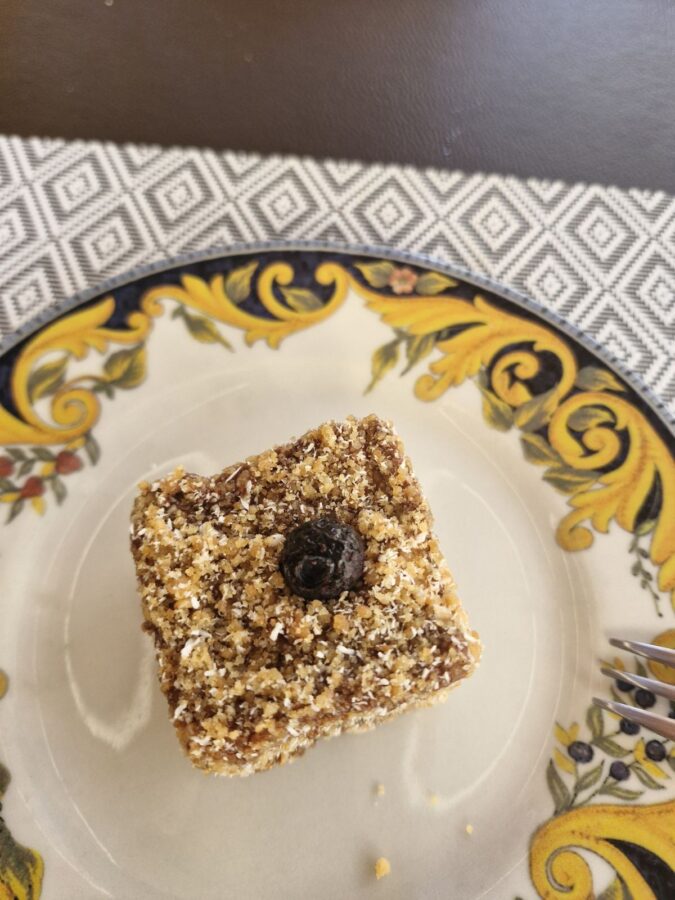 A square piece of cream-based cake topped with finely crushed nuts or biscuit crumbs and a single dark berry in the center, served on a white plate with a yellow and blue ornate border.