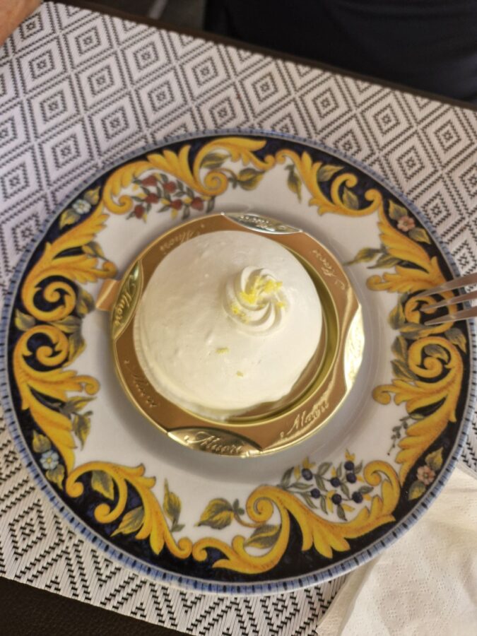 A dome-shaped Delizia al Limone dessert coated in smooth white lemon cream, finished with a small swirl and lemon zest on top, served on a gold cardboard base over a decorative blue and yellow patterned plate at La Sbirra restaurant.