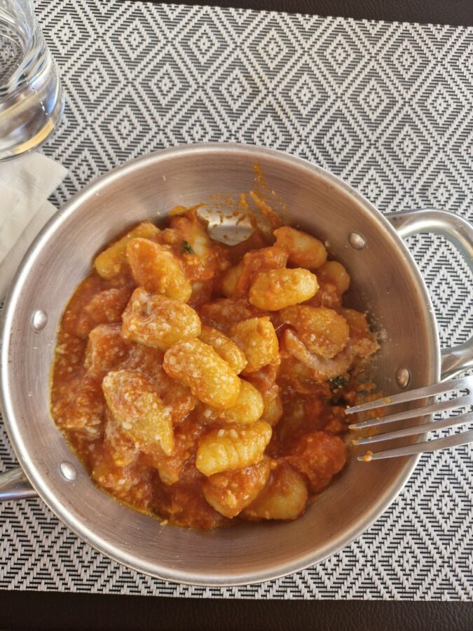 Potato gnocchi mixed with a rich tomato sauce and topped with grated cheese, served inside a small two-handled aluminum dish on a black and white geometric placemat at La Sbirra restaurant in Termoli.
