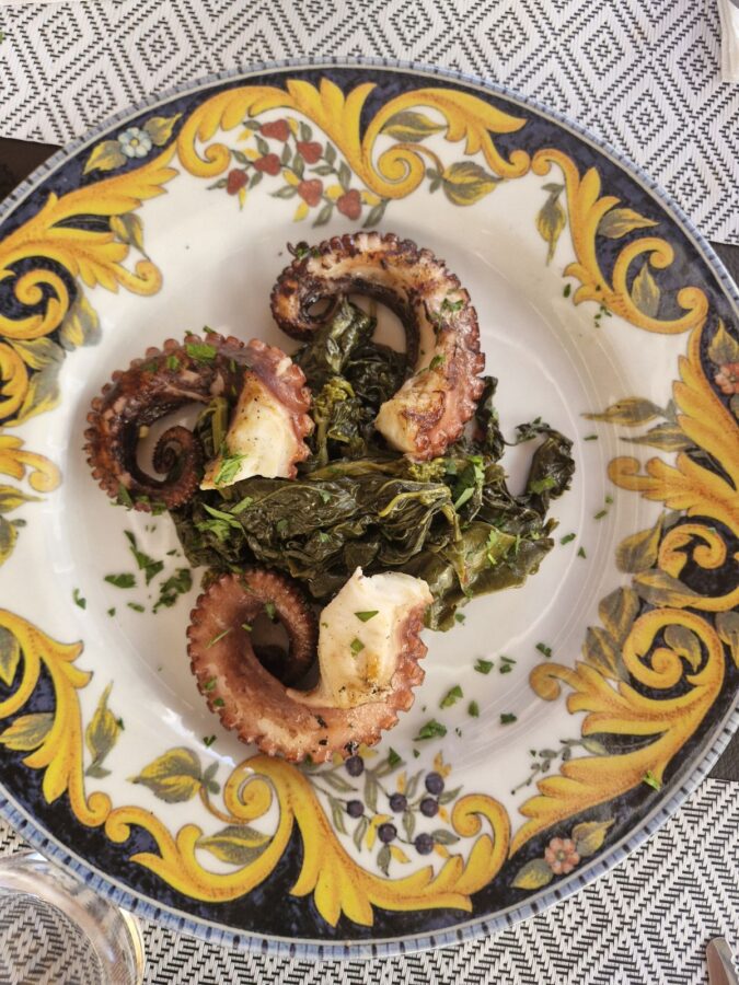 Three grilled octopus tentacles served over a bed of cooked green leafy vegetables on a decorative white plate with a prominent yellow and blue ornate border.