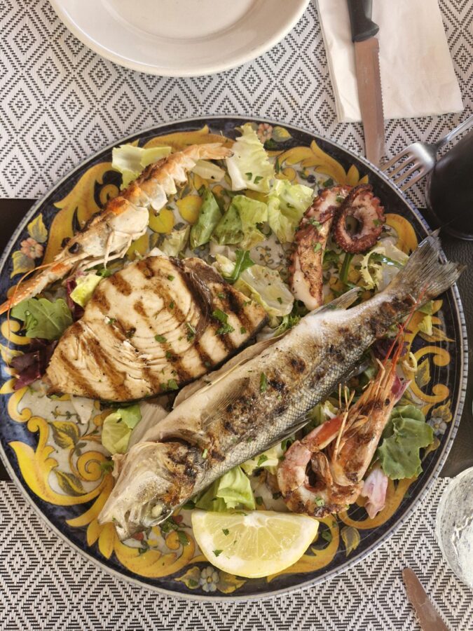 A grilled seafood plate featuring a whole white fish with grill marks, a large piece of grilled swordfish steak, an octopus tentacle, a langoustine, and a shrimp, all served over a bed of green salad with a lemon wedge on a decorative yellow and blue patterned plate.