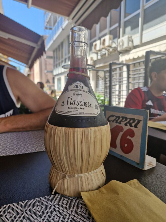 A bottle of "a Fiaschett" Falanghina IGP house wine by Fratino Cantine, served in a traditional rustic straw-covered glass fiasco bottle on an outdoor table at La Sbirra restaurant in Termoli, Molise, Italy.