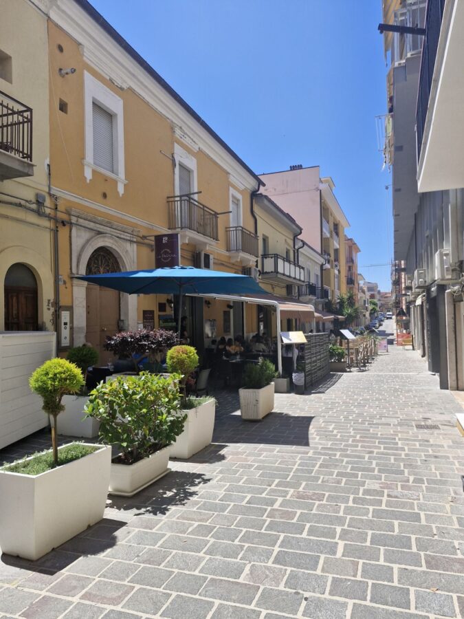 Outdoor dining area of La Sbirra restaurant on a paved pedestrian side street in Termoli, Molise, Italy, featuring a large blue umbrella, several tables with seated guests, and white rectangular planters with green bushes lined up on the left side under a clear blue sky.