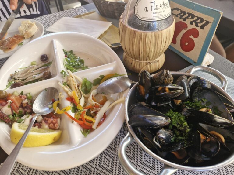 A multi-compartment white ceramic dish containing marinated anchovies, an octopus and tomato salad with lemon, and a squid salad with peppers, served next to a small metal pot filled with steamed mussels at La Sbirra restaurant. La Sbirra Termoli review.