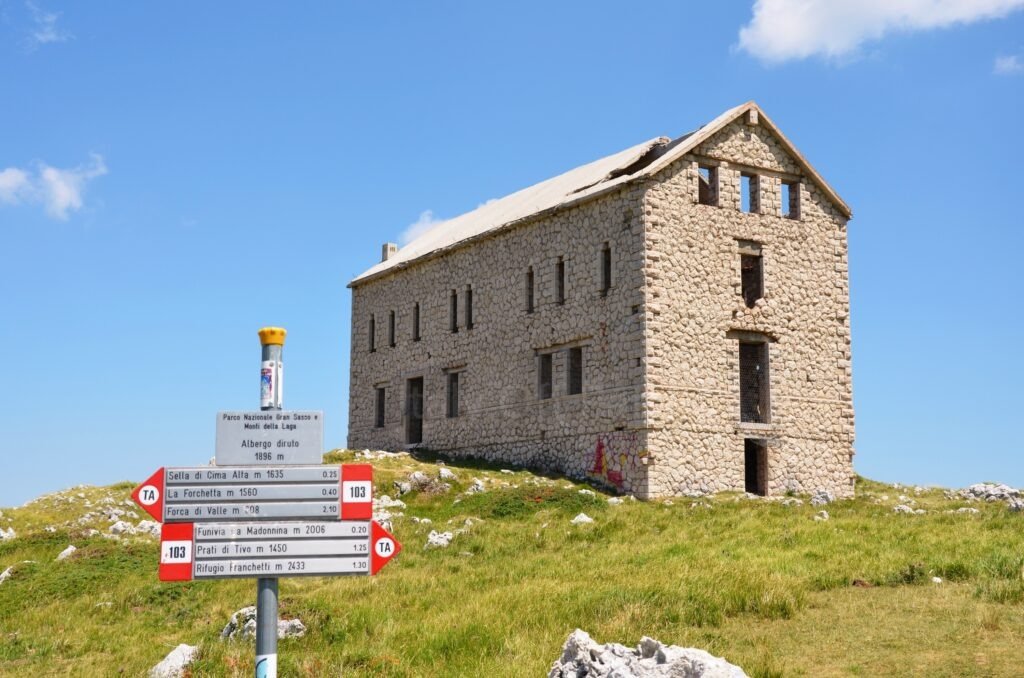 L'Albergo Diruto dell'Arapietra, an abandoned stone building at 1896 meters, next to a red and white hiking Corno Grande trail signpost in Gran Sasso, Abruzzo, Italy.