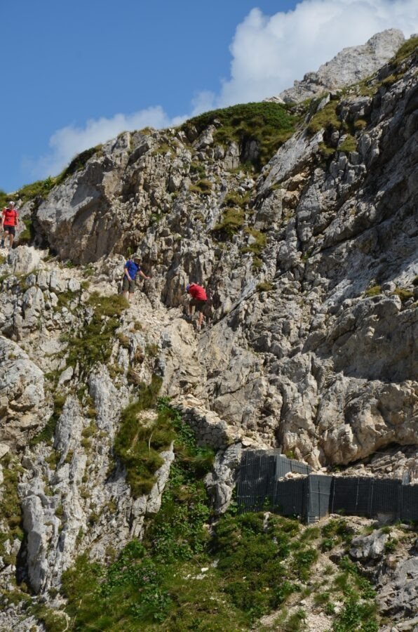 Hikers navigating a steep, rocky section secured with chains along the mountain trail to Corno Grande in Gran Sasso, Abruzzo, Italy.