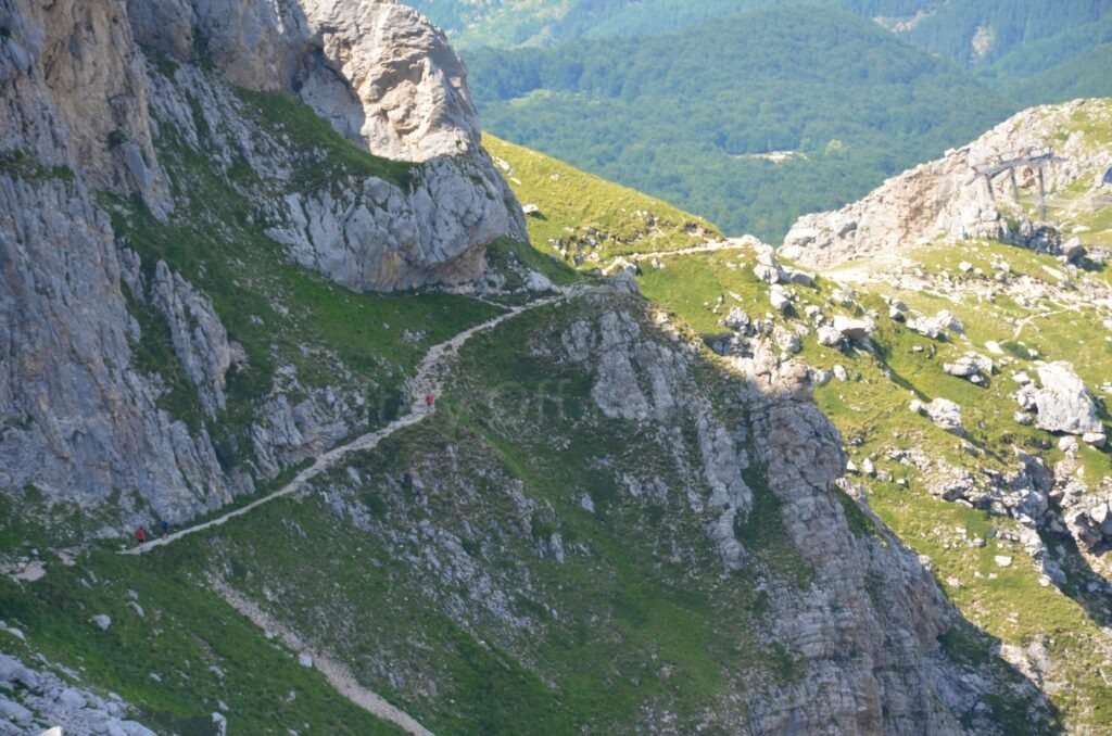 Hikers walking along a narrow, rocky mountain trail traversing a steep slope towards Corno Grande in the Gran Sasso massif, Abruzzo, Italy.