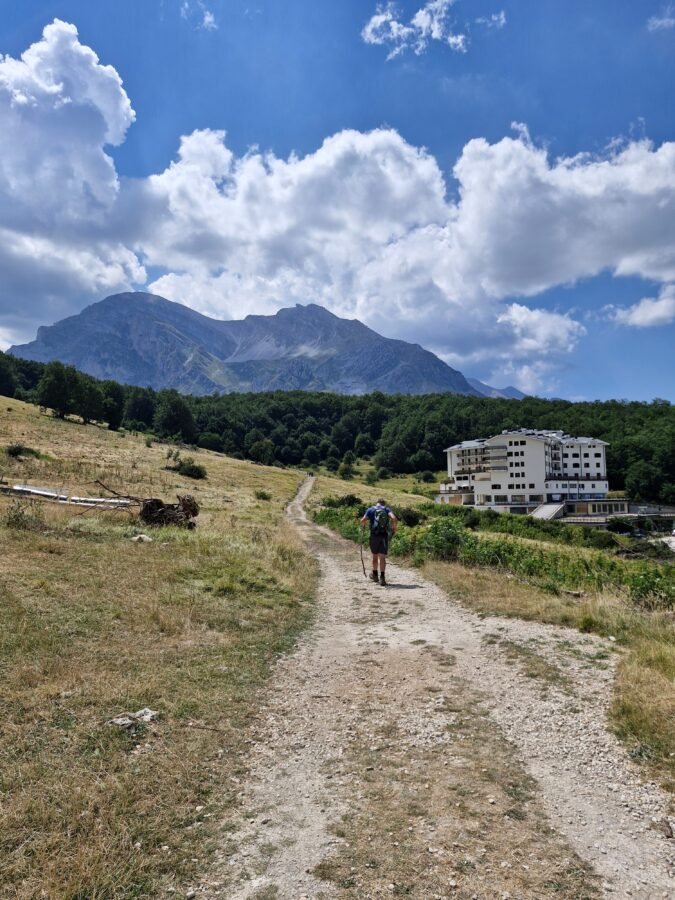 Hiker walking along the dirt path toward Val Maone with mountain slopes and hotel buildings in Prati di Tivo, Abruzzo, Italy