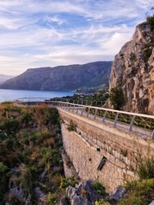 Stone skywalk pathway with metal railings built along a rocky cliffside, overlooking the sea and coastline in Maratea, Basilicata, Italy.