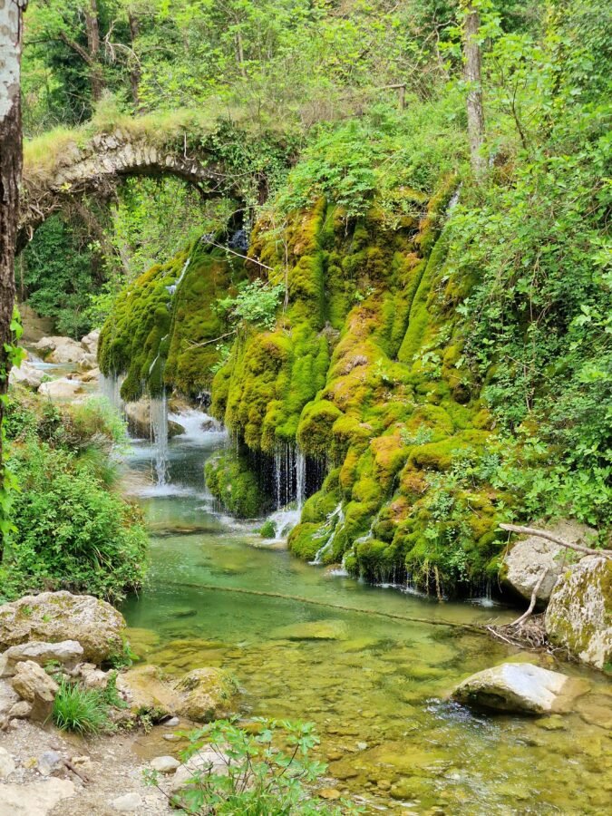 Old stone arch bridge spanning above a clear stream with moss-covered cliffs and dripping waterfalls at Capelli di Venere, Casaletto Spartano.