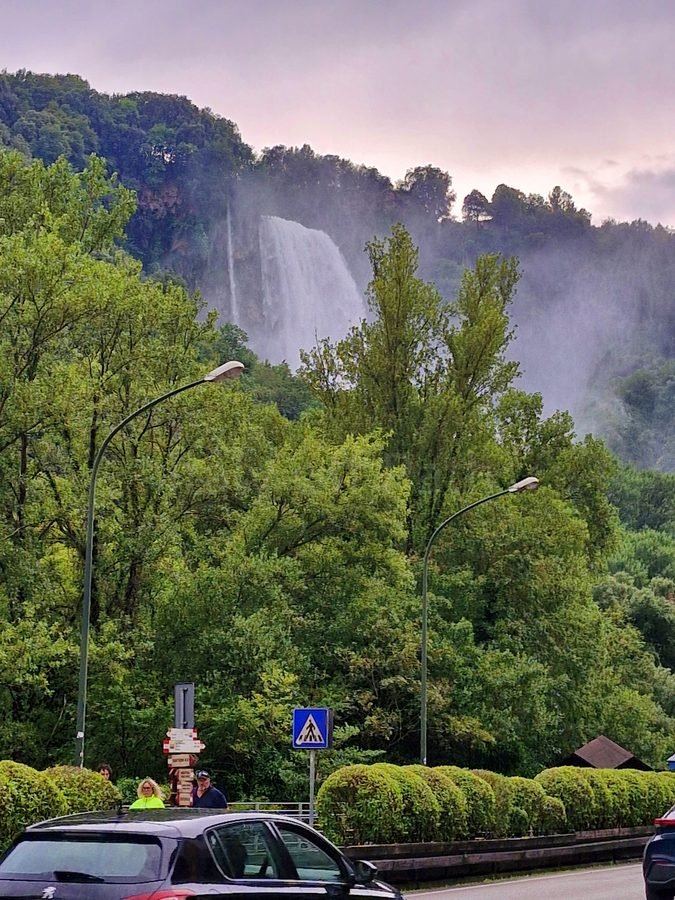 View of the upper tier of the Cascata delle Marmore waterfall shrouded in mist above dense green trees and a roadside barrier, Umbria, Italy.