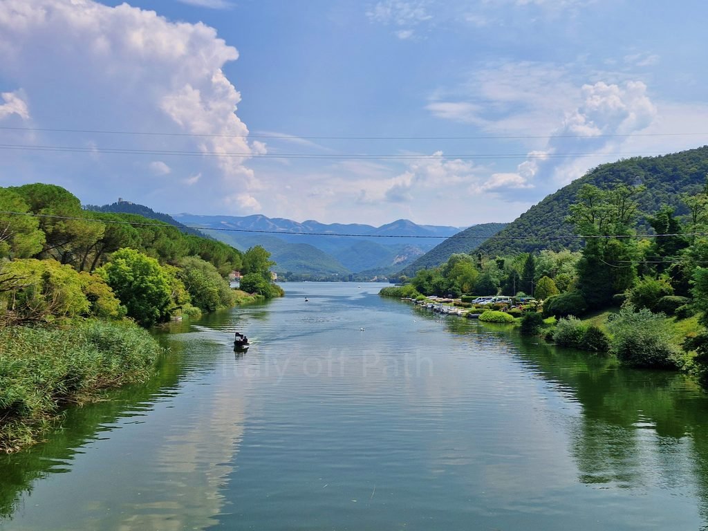 View down the calm waterway of Lake Piediluco framed by green shores and hills under a partly cloudy sky, Umbria, Italy.