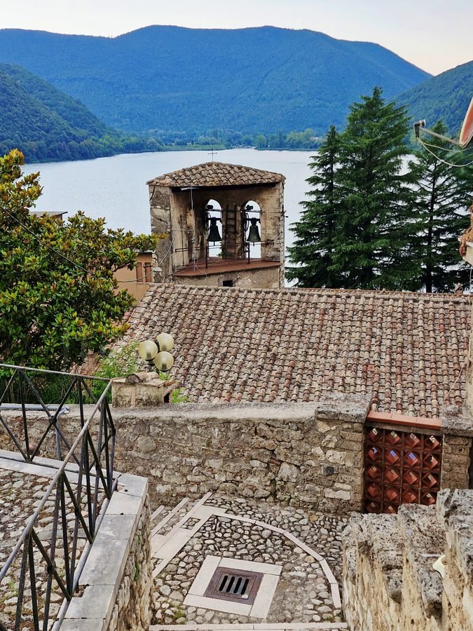 Cobblestone steps in Piediluco village leading past stone walls and terracotta roofs toward the church bell tower, with Lake Piediluco and hills behind.