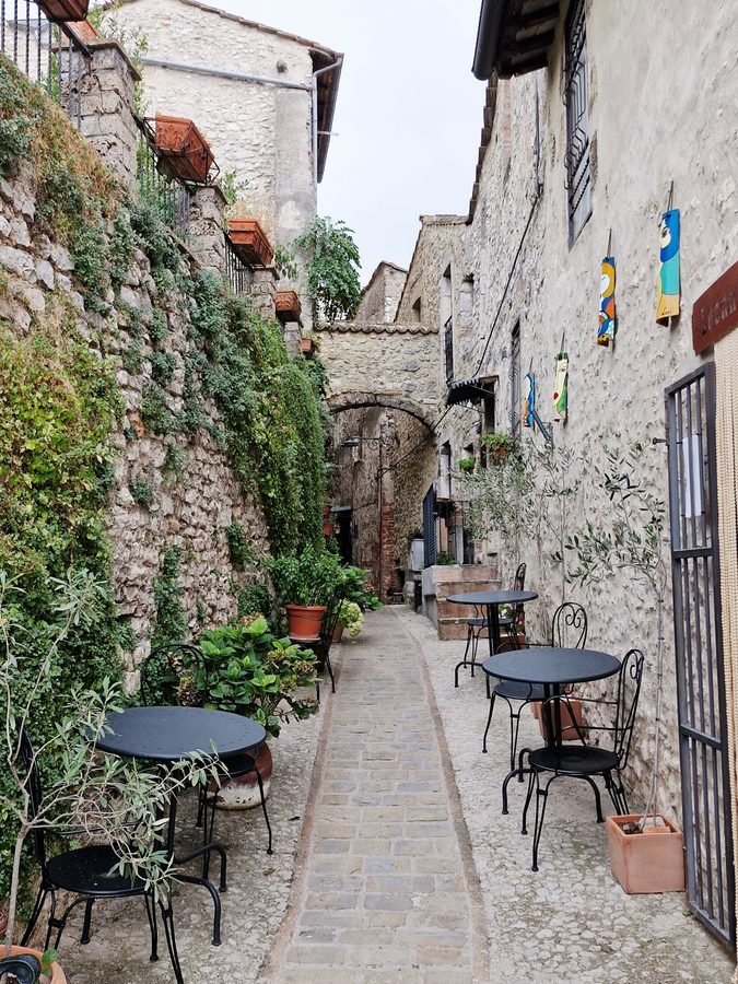 Narrow cobblestone alley in the historic center of Arrone with small cafe tables, potted plants, and a stone arch connecting the buildings, Umbria, Italy