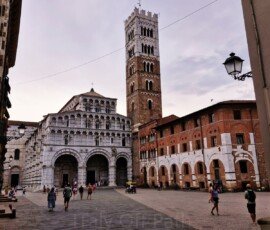 A wide shot of San Martino Cathedral in Lucca, Italy, featuring its white and grey marble facade with multiple tiers of open loggias and a tall, square stone bell tower. Pedestrians cross the paved piazza in front of the cathedral and an adjacent historic brick building on the right under a cloudy sky.
