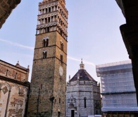 Widok kadrowany przez ciemny, kamienny łuk na Piazza del Duomo w Pistoi we Włoszech. Po lewej stronie wznosi się wysoka, kamienna dzwonnica katedry św. Zenona, obok której stoi pasiaste, marmurowe Baptysterium św. Jana. Przez szeroki, brukowany plac pod bezchmurnym, popołudniowym niebem spaceruje kilka osób, w tym jedna wyprowadzająca dwa psy.