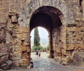 A massive, ancient Etruscan stone archway in Volterra, Tuscany, constructed from large, weathered block masonry. Through the deep arch, people walk along a paved path, leading to a view of a single tall cypress tree and distant, hazy Tuscan hills.
